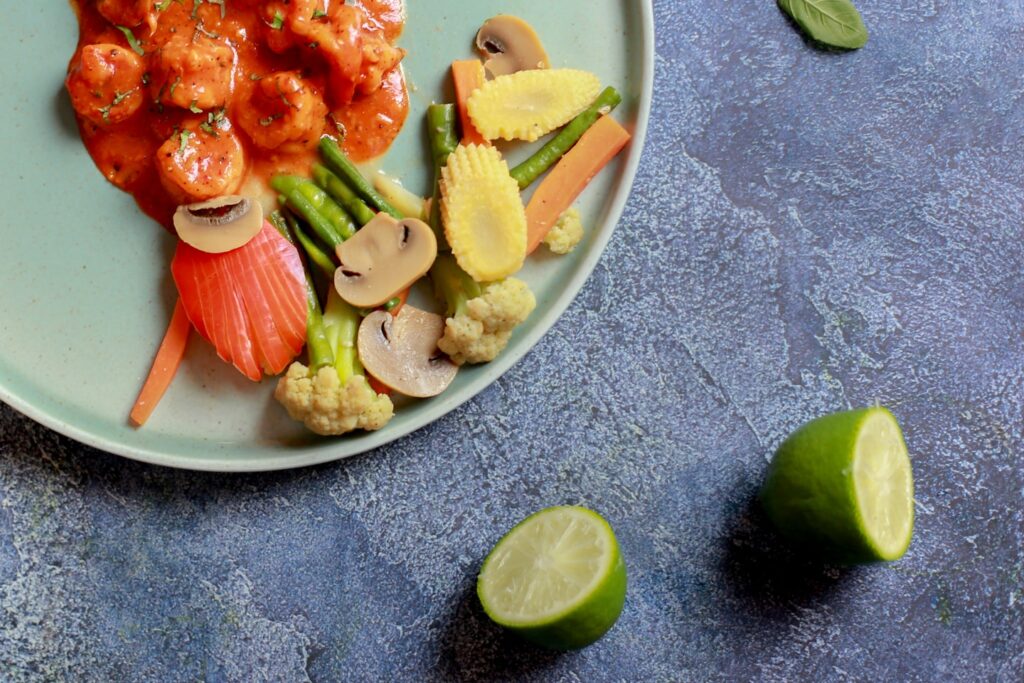 An overhead shot of A vibrant top-down shot of shrimp in a red sauce next to perfectly blanched vegetables (baby corn, mushrooms) on a light blue plate, garnished with lime and basil.